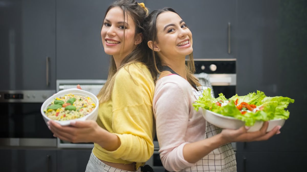Two smiling women hold vibrant salads in a kitchen setting, showcasing fresh culinary creations.